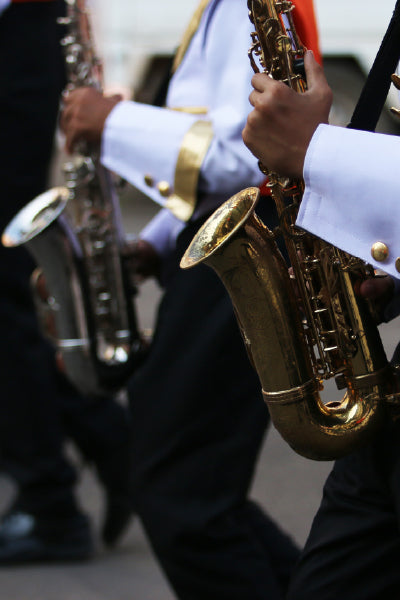 Close-up of marching band members in uniform playing saxophones during a parade. The musicians wear crisp white jackets with gold trim and dark pants, and their instruments gleam in the light as they march in step on a paved street.