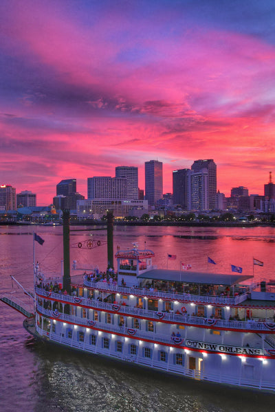 A classic riverboat named "City of New Orleans" glides along the Mississippi River at sunset, with the New Orleans skyline in the background. The sky is ablaze with vibrant pink, purple, and orange hues, reflecting off the water and casting a warm glow over the cityscape.