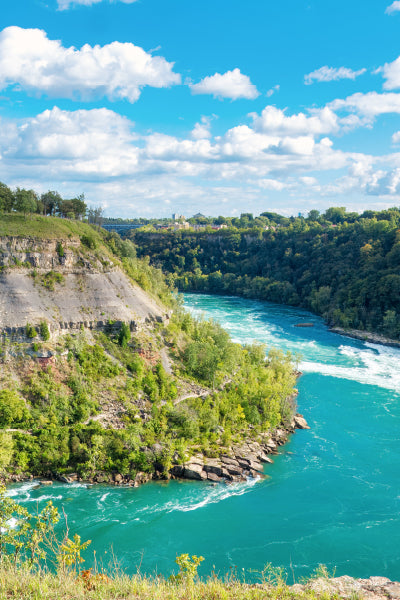 A scenic view of the Niagara River winding through a lush, green gorge with steep rocky cliffs on one side and dense forest on the other, under a bright blue sky dotted with fluffy white clouds.