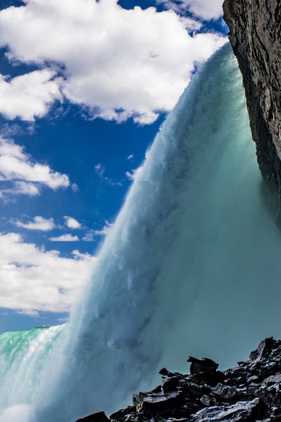 A striking side-angle view from behind a massive waterfall, showing a thick, powerful curtain of turquoise water plunging downward past a rocky cliff face. The sky above is bright blue, filled with scattered fluffy white clouds. In the foreground, jagged dark rocks line the bottom right corner, emphasizing the raw natural force of the scene.