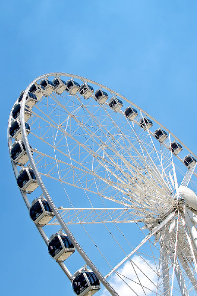 An upward view of a large Ferris wheel against a clear blue sky. The white metal structure is detailed with spokes and features enclosed, black-and-white passenger cabins evenly spaced around the wheel. The bright daylight casts shadows and highlights the geometric lines of the Ferris wheel.