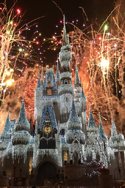 A dazzling night scene of Cinderellaβs Castle at Walt Disney World, covered in sparkling white holiday lights, with vibrant fireworks bursting in the sky behind it.