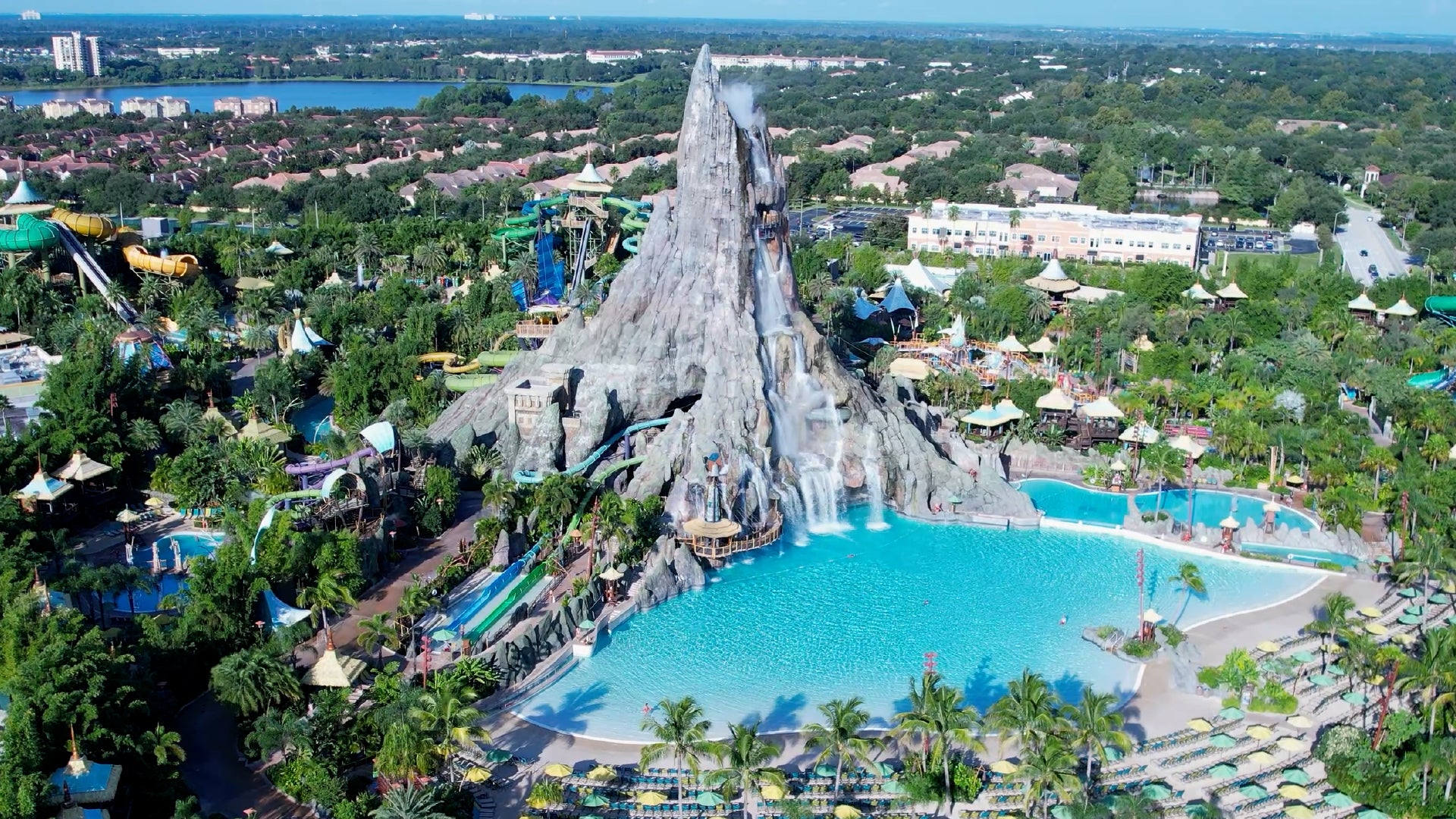An aerial view of a tropical water park featuring a towering artificial volcano with cascading waterfalls flowing into a large turquoise wave pool. Surrounding the volcano are winding water slides, lush greenery, and palm trees, creating a paradise-like atmosphere. The park is filled with various attractions, including shaded cabanas, pools, and thrilling rides, while a nearby residential area and lake are visible in the background.