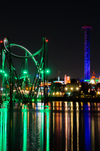 A vibrant night view of a theme park with a brightly lit roller coaster featuring green lights, tall thrill rides illuminated in blue and purple, and their colorful reflections shimmering on the water below.