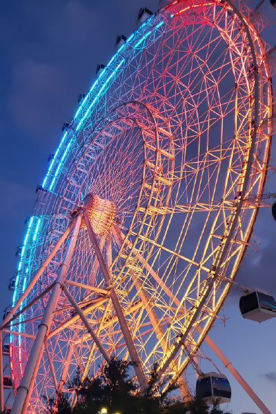 A large Ferris wheel illuminated with colorful lights in shades of blue, pink, and yellow against a dusk sky, with gondola cabins visible around the rim.
