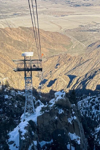Aerial view of a snow-covered mountain landscape with a tramway tower and cables extending down into a desert valley. The image shows the transition from the rugged, forested mountains in the foreground to the arid, flat terrain below, dotted with wind turbines and a small settlement.