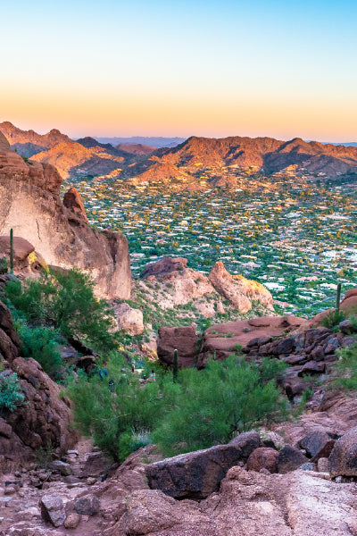 Scenic view from Camelback Mountain in Phoenix, Arizona, showing a rocky desert trail with cacti and green shrubs in the foreground, and a sprawling suburban cityscape below, surrounded by rugged mountains under a colorful sunset sky.