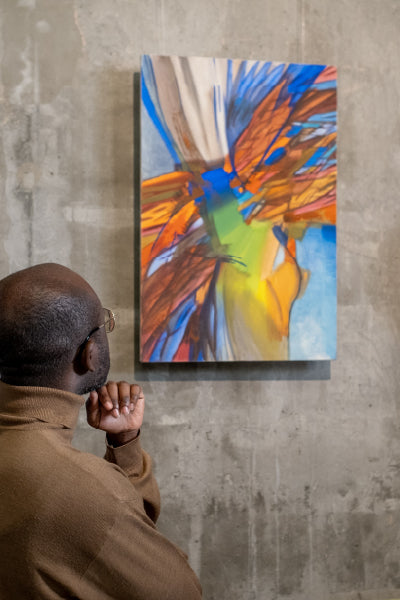 Man wearing glasses and a brown turtleneck thoughtfully observes a colorful abstract painting mounted on a concrete wall, featuring dynamic brushstrokes in blue, orange, yellow, and green tones.