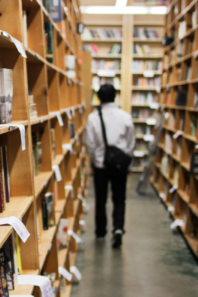 Person with a shoulder bag walking down a narrow aisle between tall wooden bookshelves in a library or bookstore, with books and category labels lining the shelves and a softly blurred background.
