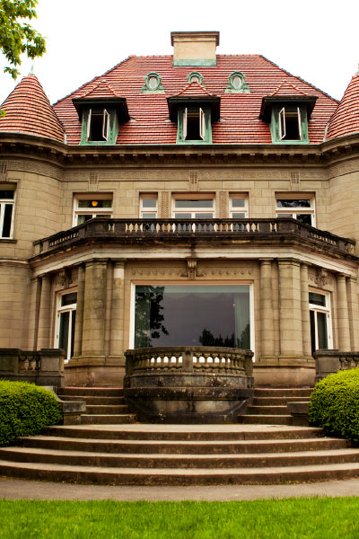 Front view of a grand historic mansion with a red tiled roof, ornate stone facade, symmetrical windows, and a central stone staircase leading up to a columned entrance, surrounded by manicured green bushes and lawn.