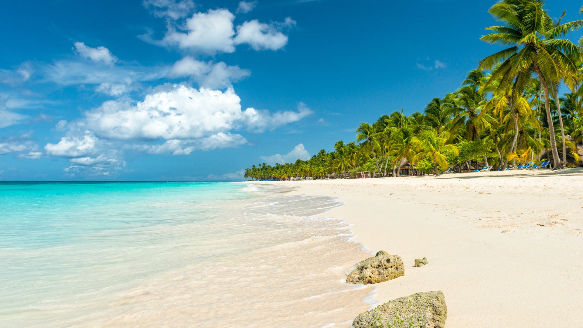 A tropical beach with soft white sand, turquoise waters, and gentle waves washing ashore. The coastline is lined with lush green palm trees, and a few rocks are scattered on the sand. The sky is bright blue with fluffy white clouds, creating a serene and picturesque paradise.