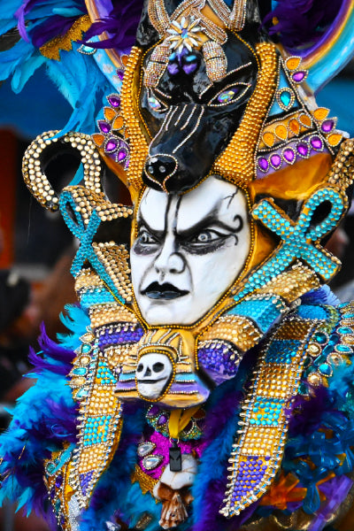 Person wearing an elaborate and colorful costume with blue feathers, gold and purple beading, and intricate designs including an Anubis-like black headdress and a white make painted with dramatic black patterns and dark lips.