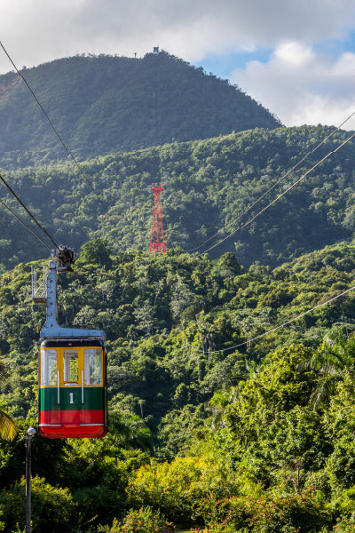 Colorful cable car suspended on a cable line above a lush green tropical forest, with a red support tower in the distance and a large forested mountain in the background under a partly cloudy sky.