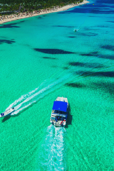 Aerial view of two boats cruising on clear turquoise water near a beach lined with palm trees and umbrellas. The larger boat has a blue roof and leaves a white trail behind, while a smaller speedboat follows alongside, also creating a wake in the water. The vibrant blue ocean contrasts with the sandy shore and green foliage in the background.