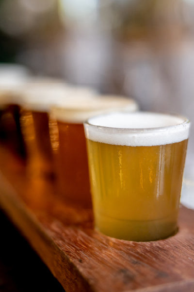 A close-up view of a flight of craft beers served in small glasses on a wooden board, with a frothy head on each beer. The beers vary in color from pale to amber, and the background is softly blurred.