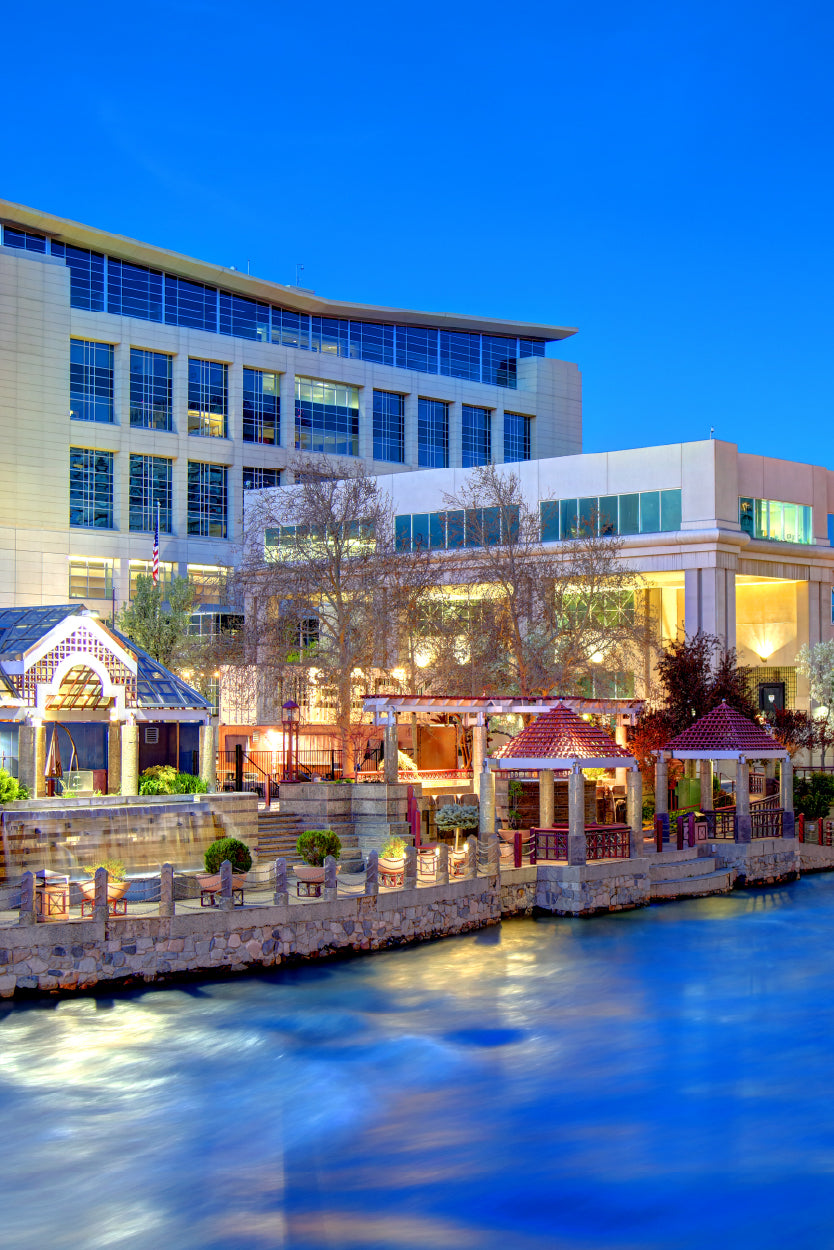 A vibrant scene along the Truckee Riverwalk. The brightly lit buildings, walkways, and pavilions by the river create a lively atmosphere, especially with the reflection of lights on the water during twilight. The architecture and waterfront setting are characteristic of this popular urban destination known for dining, shopping, and cultural attractions.