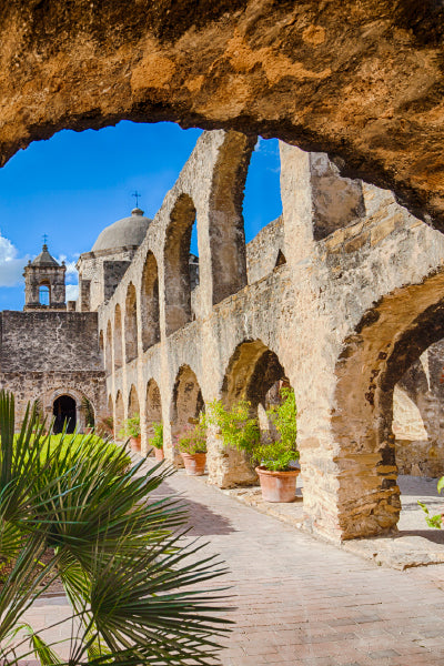 Historic stone arches and walls of an old mission or colonial building, with potted plants lined along the walkway. The architecture features multiple rounded arches and a domed roof in the background under a bright blue sky. Green palm fronds are visible in the foreground.