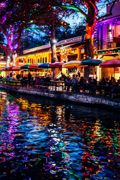 A vibrant riverside scene at night with colorful string lights wrapped around trees and buildings. People are sitting under umbrellas along the water's edge, enjoying the lively atmosphere. The water reflects the bright, multicolored lights, creating a beautiful shimmering effect.
