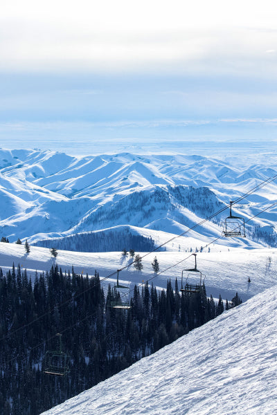 A scenic winter landscape with snow-covered mountains under a pale blue sky. In the foreground, a ski slope descends with tall evergreen trees at the base. Several empty ski lift chairs are suspended on cables, stretching across the slope toward the mountain peaks in the distance.