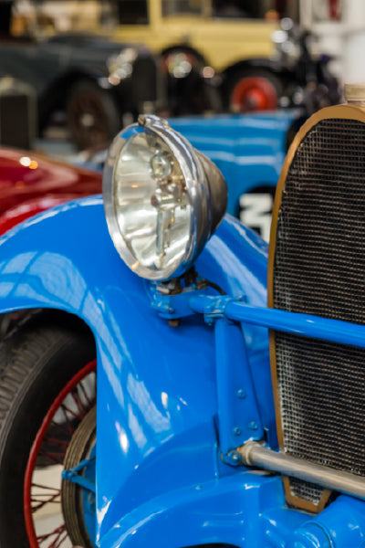 Close-up of the front of a vintage blue car, focusing on the round headlight mounted on the curved fender and part of the grille. In the background, other classic cars are partially visible and out of focus.