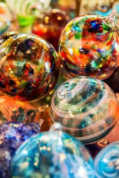 A close-up view of a vibrant collection of hand-blown glass ornaments, each uniquely decorated with swirling patterns and a variety of bright colors including red, blue, green, orange, and purple. The smooth, glossy surfaces reflect light, highlighting the intricate craftsmanship and creating a festive, artistic atmosphere.