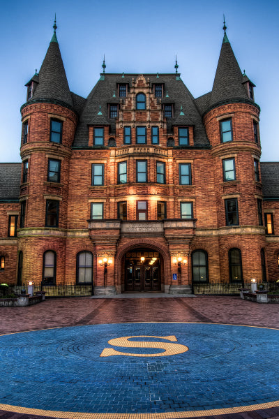 A large, historic brick building with multiple windows and two tall, rounded towers on either side. The building has a steep, dark slate roof with decorative spires. In front of the entrance is a circular blue brick area with a large yellow letter "S" in the center. The building is illuminated by warm exterior lights at dusk.