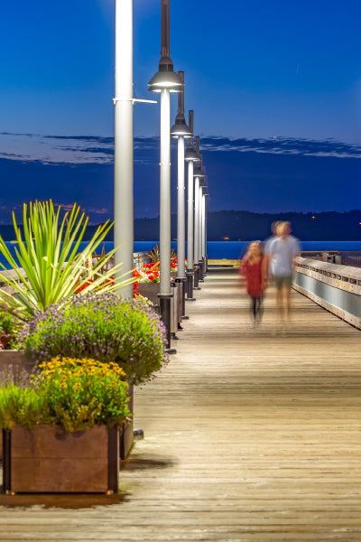 A wooden pier at dusk, lined with tall lamp posts and colorful flower planters on the left side. Two blurred figures, likely a child and an adult, are walking away from the camera towards the water. The sky is deep blue with faint clouds and distant land visible on the horizon.