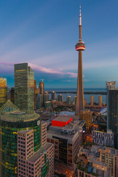A cityscape at dusk featuring the iconic CN Tower standing tall among modern skyscrapers. The buildings are illuminated by the fading sunlight and city lights, with a large body of water visible in the background under a clear sky.