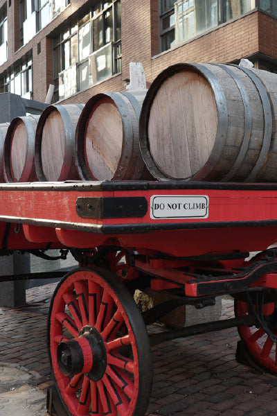 A vintage red wooden cart with large spoked wheels holding several wooden barrels. The cart has a sign that reads "DO NOT CLIMB" and is parked on a cobblestone street in front of a brick building with large windows.