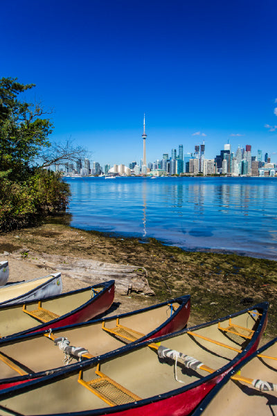 Several red and tan canoes are lined up on the shore of a calm body of water, with the Toronto skyline and CN Tower clearly visible across the water under a bright blue sky. Trees frame the left edge of the image.