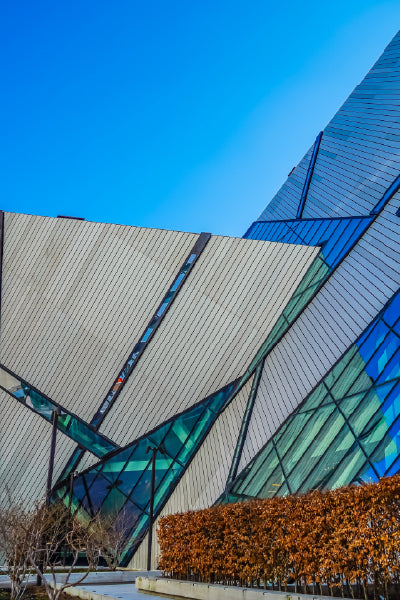 Modern architectural building with sharp, angular geometric shapes made of light-colored panels and large glass windows reflecting the blue sky. The building is surrounded by neatly trimmed brown bushes and a few leafless trees under a clear, bright blue sky.