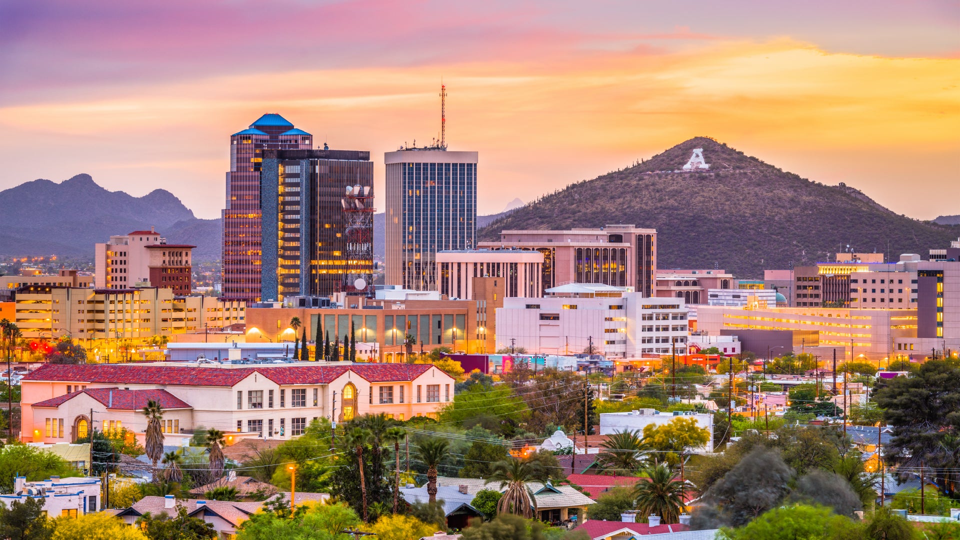 A vibrant cityscape of Tucson, Arizona, at sunset, featuring modern high-rise buildings alongside historic red-roofed buildings in the foreground. The skyline is backed by rolling desert mountains, with the iconic "A" on Sentinel Peak visible. The sky is painted in warm hues of pink, orange, and purple, casting a golden glow over the city, while palm trees and lush greenery add a touch of nature to the urban landscape.