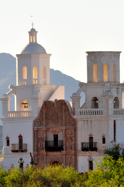 A historic white Spanish colonial-style mission with two bell towers and intricate architectural details, set against a backdrop of mountains and bathed in warm sunlight.