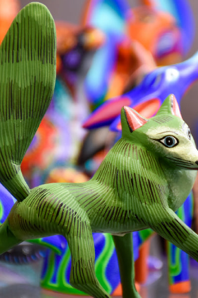 Close-up of a brightly colored, green wooden animal figurine with large eyes and a bushy tail, part of a collection of traditional Mexican alebrijes with vivid, multicolored patterns in the background.
