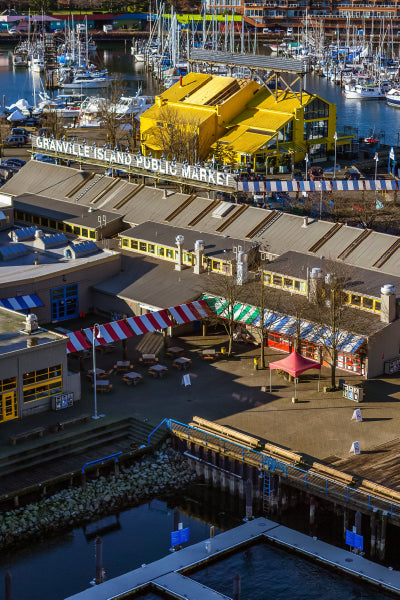 Aerial view of Granville Island Public Market in Vancouver, featuring colorful market buildings with red, blue, and white striped awnings, outdoor seating areas, and a bright yellow building in the background surrounded by a marina filled with boats.