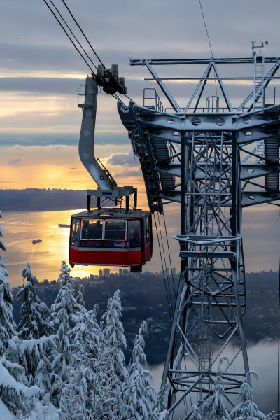 A red cable car suspended on cables glides past a tall metal tower covered in snow. Snow-covered trees are visible below, and a scenic view of water and distant land is seen in the background under a cloudy sky during sunset.