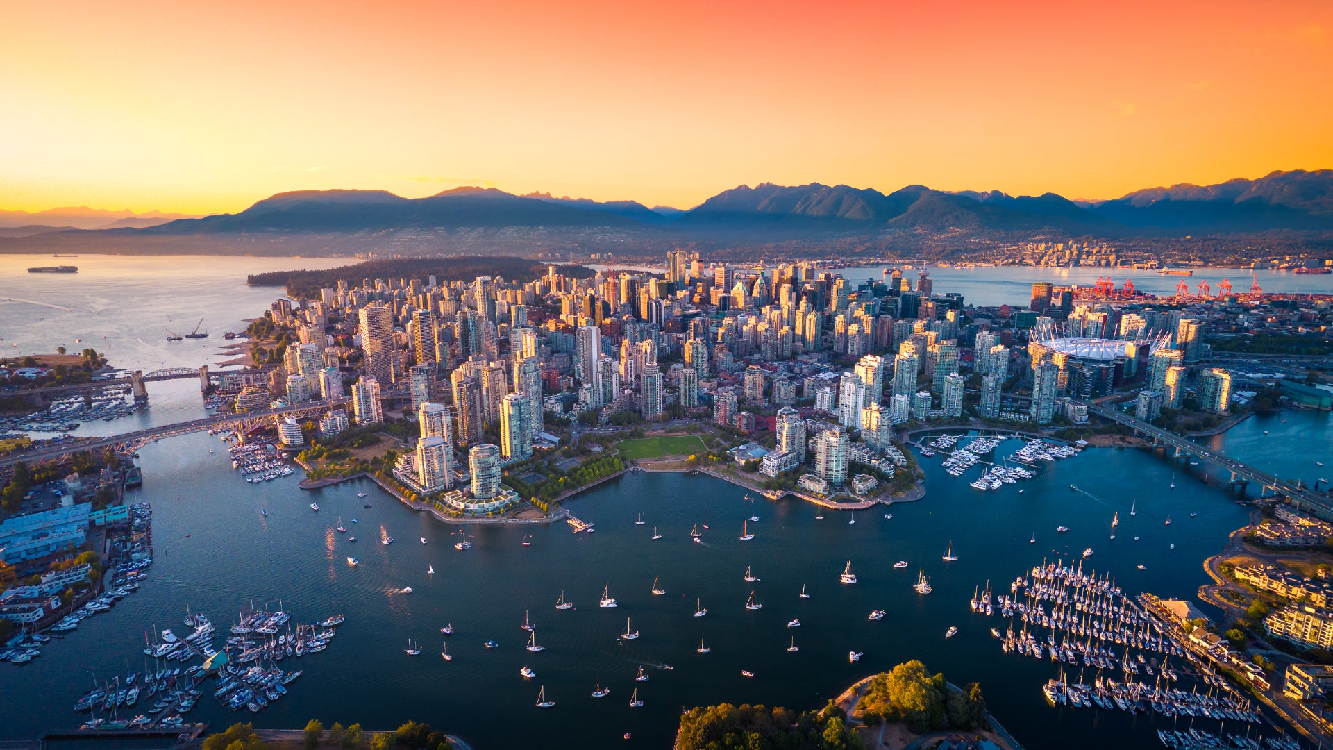 A breathtaking aerial view of Vancouver, Canada, at sunset, showcasing the city's modern skyline with high-rise buildings, a scenic harbor filled with sailboats, and the surrounding mountains in the background. The warm hues of the sunset contrast with the cool blue waters, highlighting the beauty of the urban landscape and natural scenery.