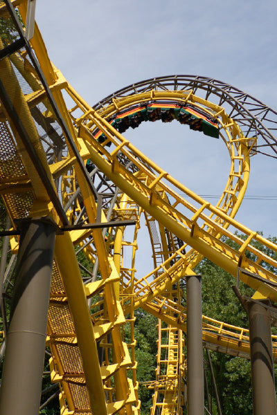 A vibrant yellow roller coaster track with multiple loops and twists, captured from a low angle. A roller coaster train full of riders is upside down in the middle of a loop, surrounded by green trees and a clear blue sky in the background.