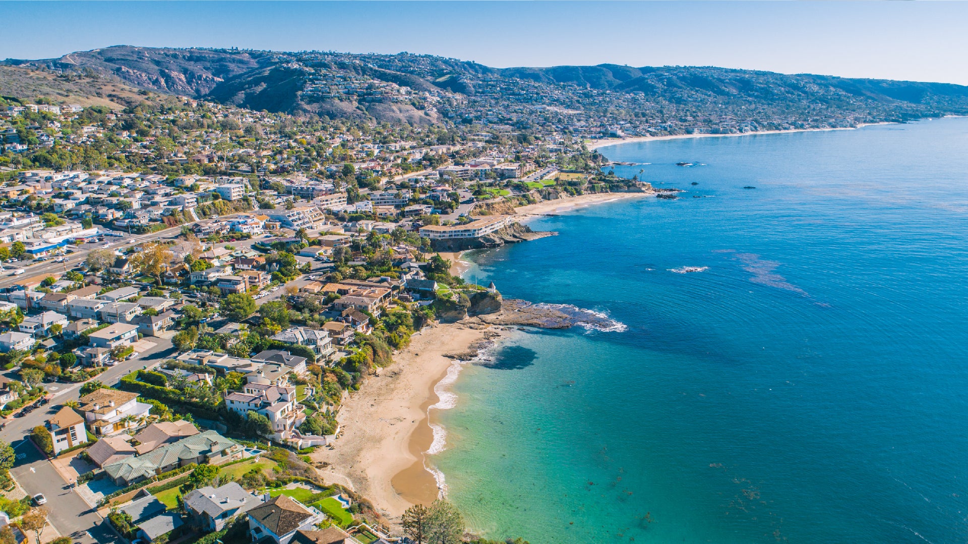A stunning aerial view of a coastal town with houses densely packed along the hillsides and shoreline, leading down to small, sandy beaches and rocky coves. The turquoise waters of the ocean are calm and clear, gently meeting the shore. Rolling hills rise in the background, covered in homes and greenery under a bright, cloudless blue sky.