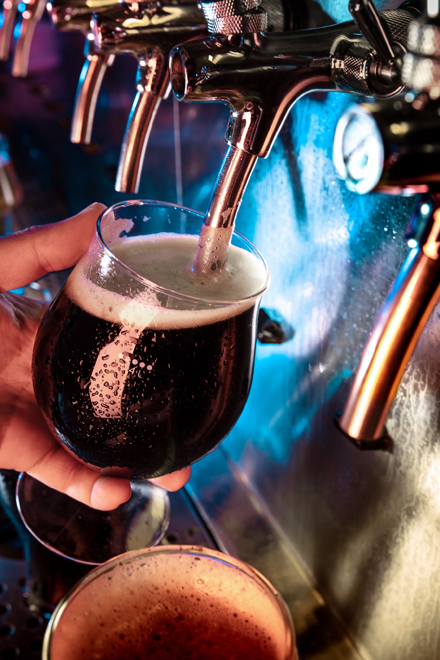 A close-up view of a person pouring a dark beer from a tap into a rounded glass at a bar, with vibrant lighting in shades of blue and purple reflecting off the metal taps and glass surfaces.