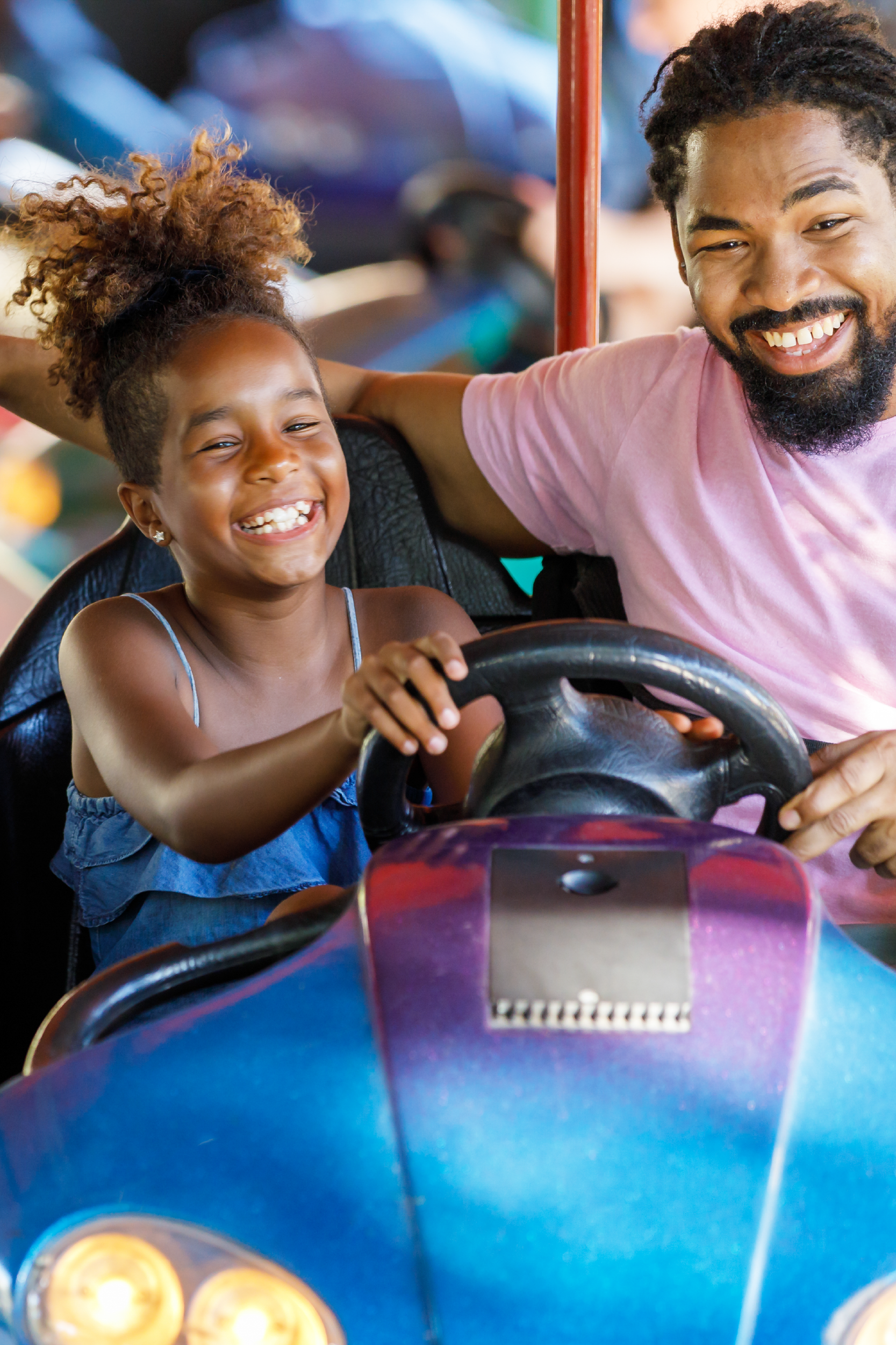 A joyful moment of a young girl and a man, possibly her father, laughing together as they ride in a bumper car at an amusement park. The girl is gripping the steering wheel with excitement, while the man sits beside her smiling brightly. The vibrant colors of the bumper car and the lively atmosphere highlight the fun and playful experience they're sharing.