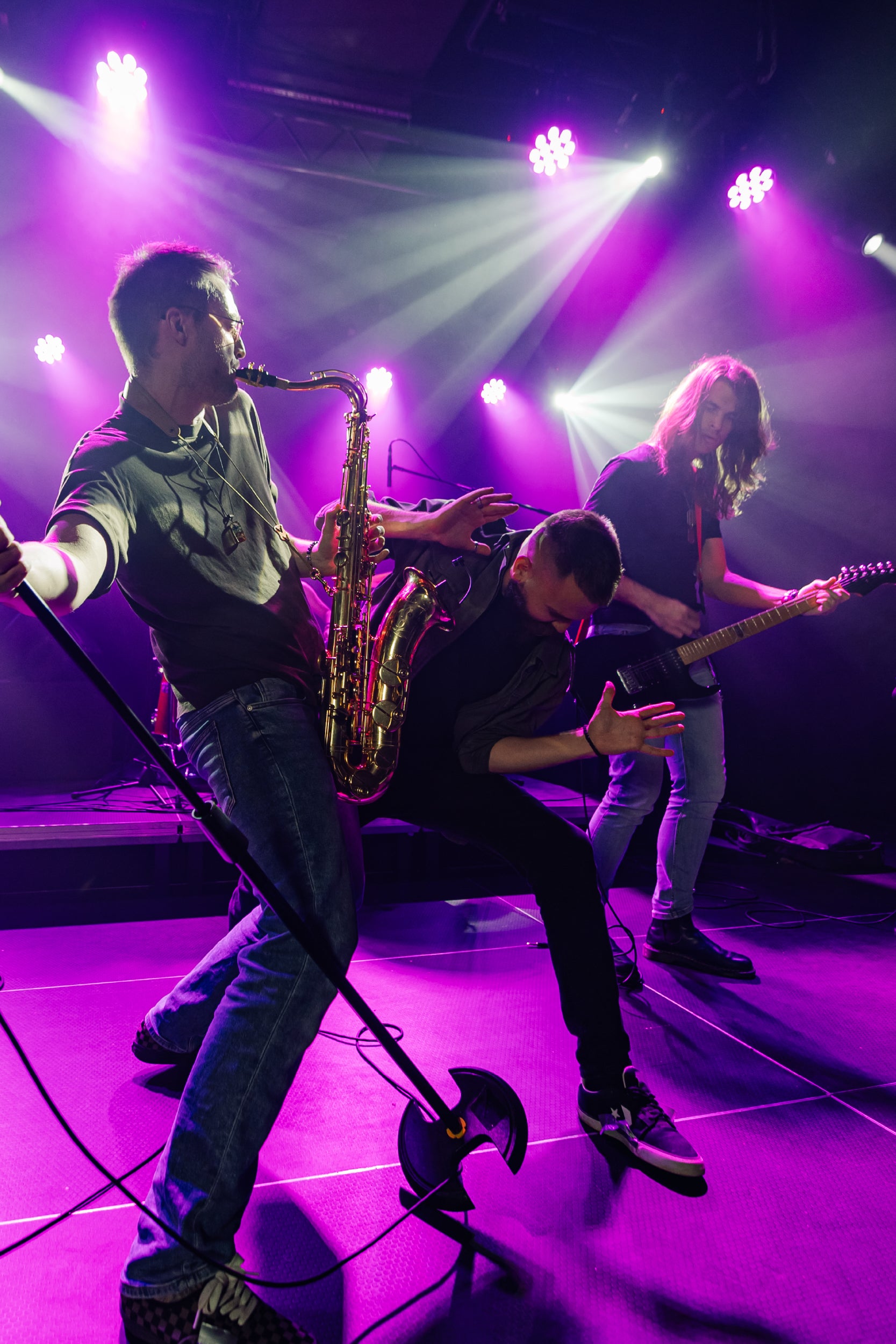 A dynamic scene of a live music performance featuring three male musicians on stage under vibrant purple lighting—one playing a saxophone, another playing an electric guitar, and the third striking a dramatic pose in front of the spotlight.