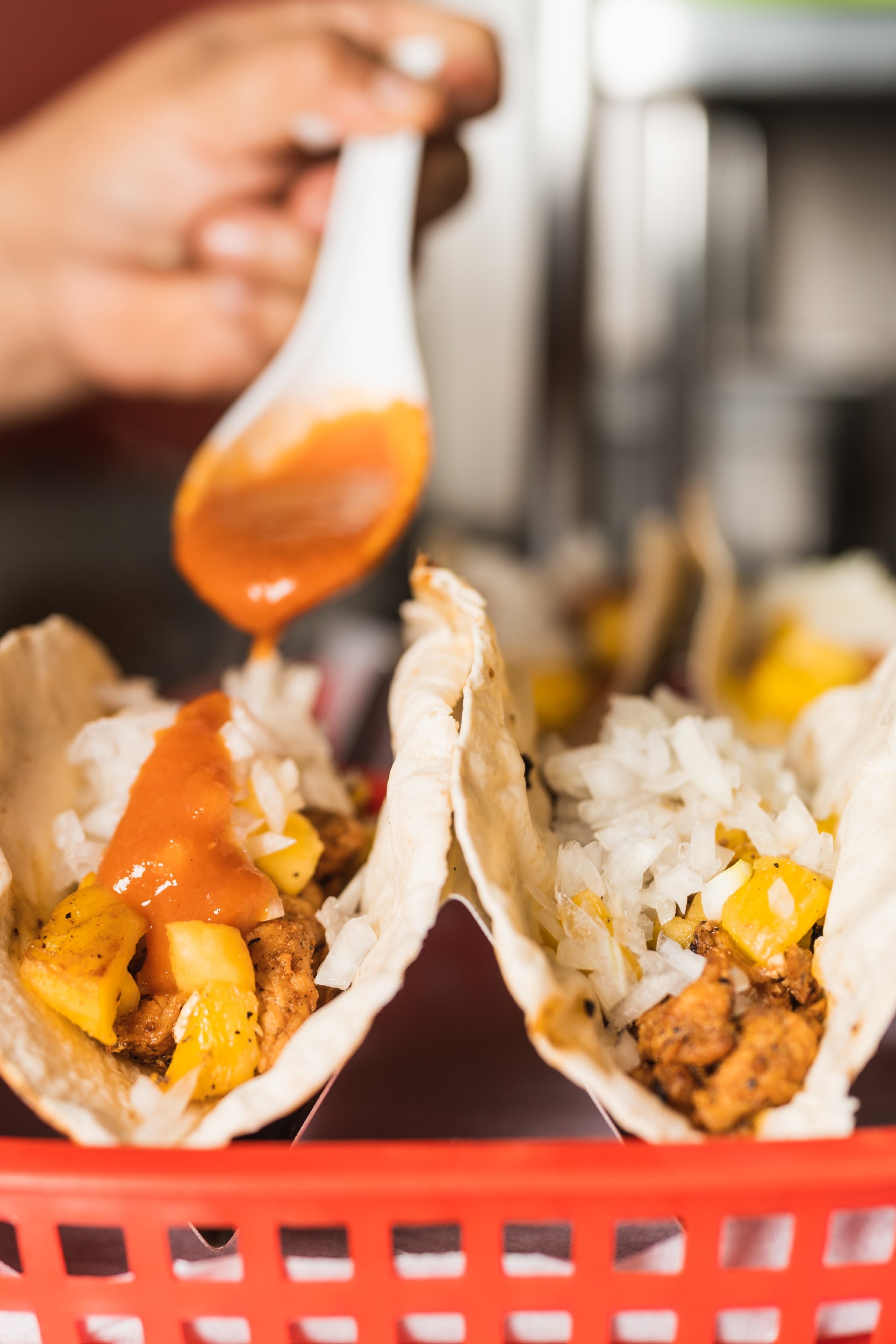 A close-up of a person spooning red sauce onto tacos filled with seasoned meat, chopped onions, and pineapple chunks, served in a red plastic basket.