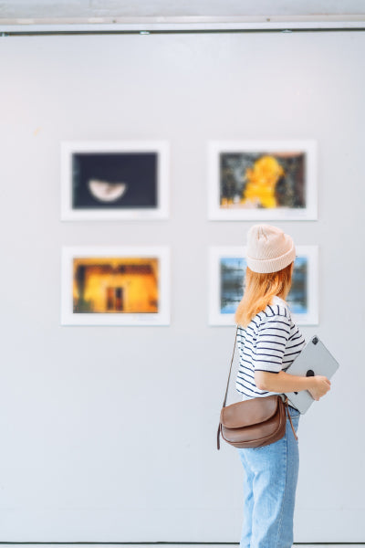 A young woman wearing a striped shirt, jeans, and a beanie stands in front of a gallery wall, holding a tablet and looking at a display of four framed photographs.