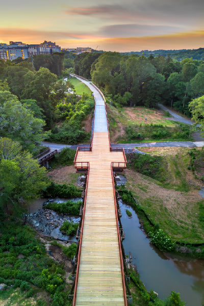 A scenic aerial view of a long wooden pedestrian bridge stretching over a stream and through lush greenery, with a sunset sky in the background and city buildings visible in the distance.