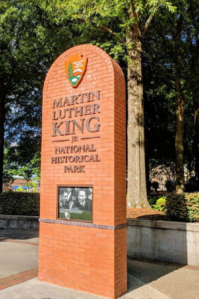 A tall brick monument sign marking the entrance to the Martin Luther King Jr. National Historical Park, featuring the National Park Service emblem and a black-and-white photo of Dr. King, set against a backdrop of trees and a sunny day.