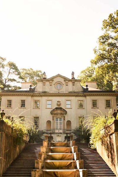 A grand historic mansion with classical architecture, viewed from the base of a dramatic stone staircase lined with greenery, bathed in warm sunlight and surrounded by tall trees.