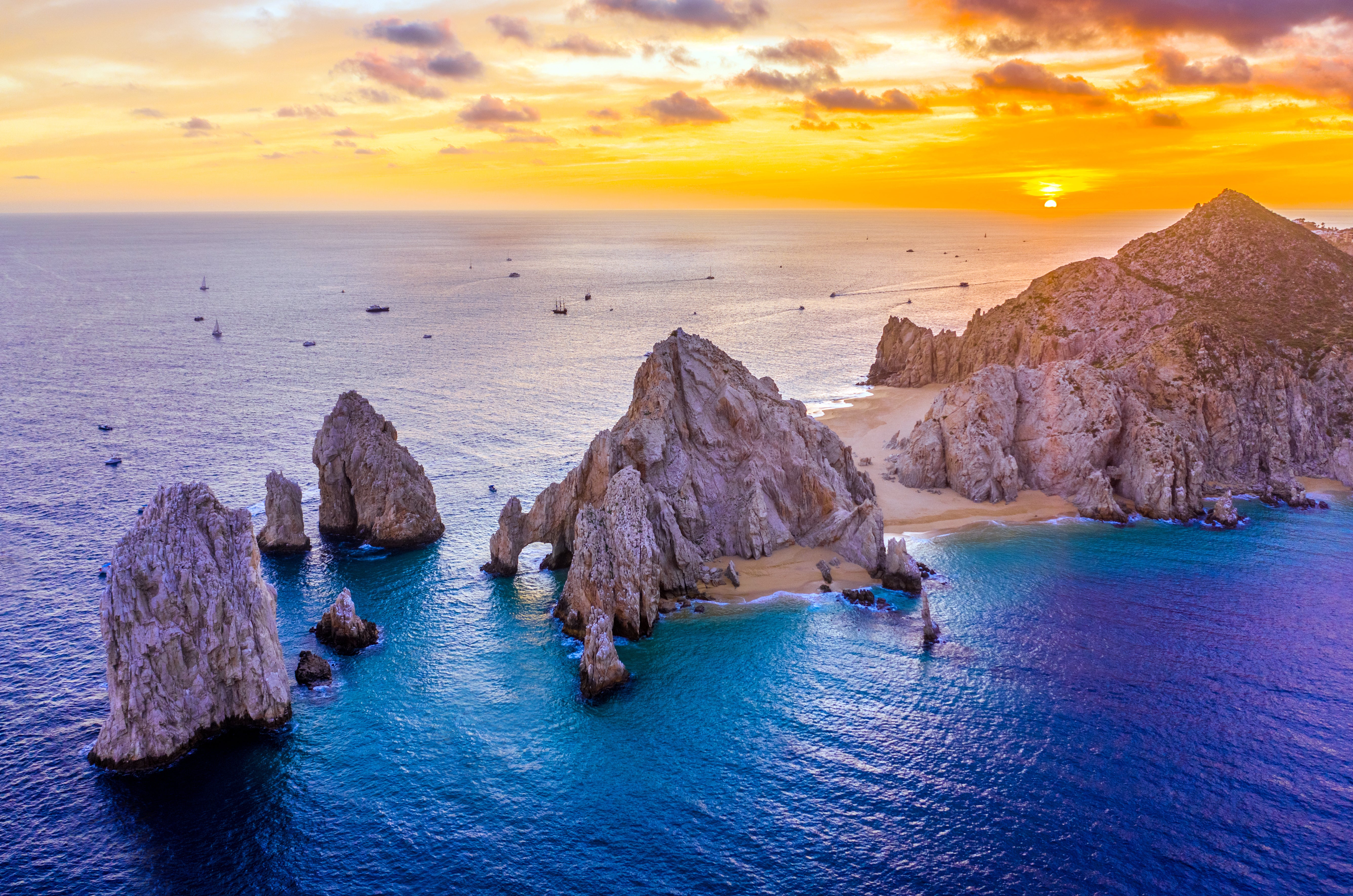 A breathtaking aerial view of El Arco in Cabo San Lucas, Mexico, captured at sunset. The iconic rock formations rise dramatically from the turquoise waters where the Pacific Ocean meets the Sea of Cortez. Boats are scattered across the horizon, and golden sands of Lover’s Beach and Divorce Beach contrast beautifully with the rugged cliffs. The sky is painted with warm orange and purple hues as the sun dips below the horizon.