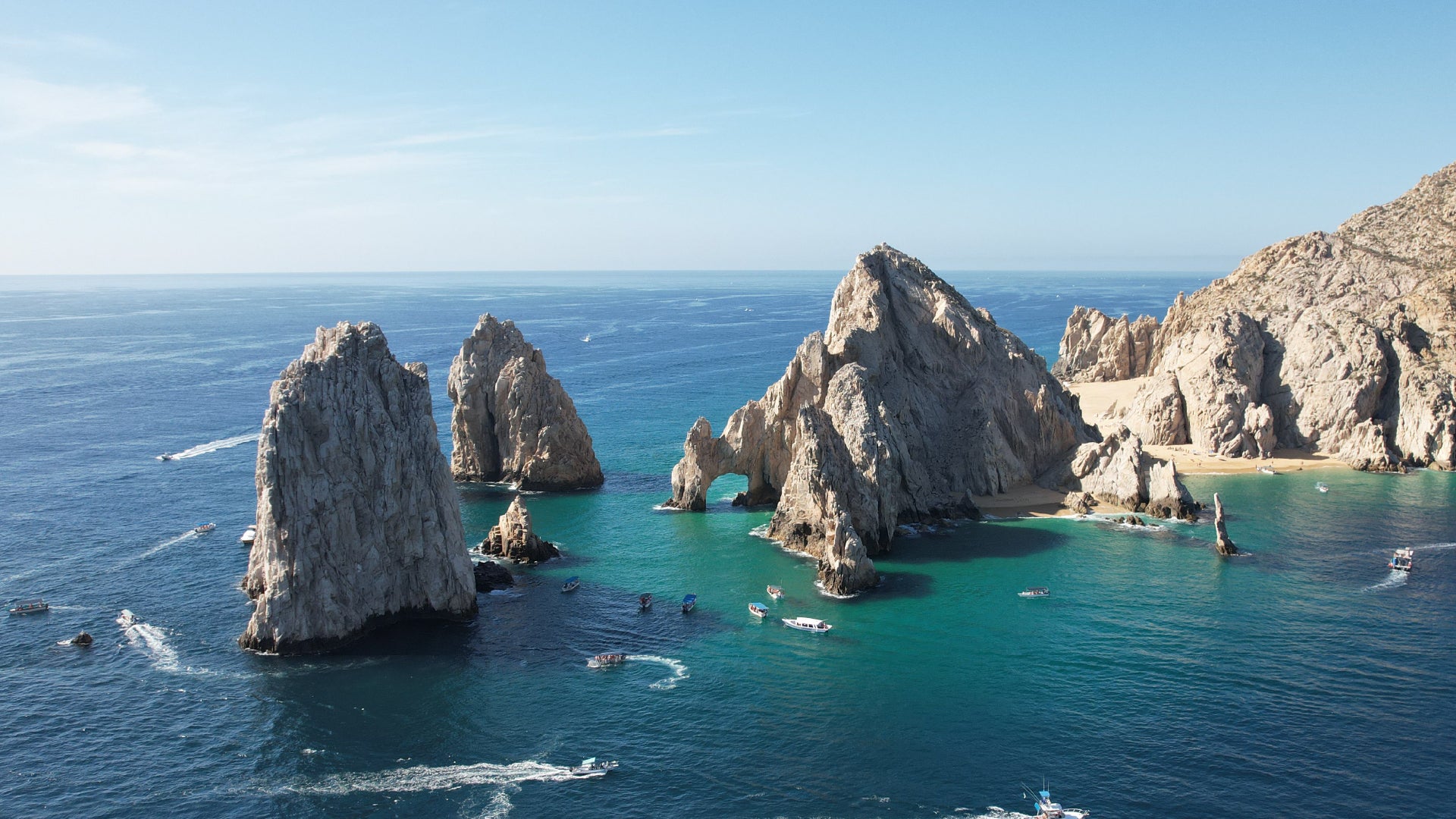 A breathtaking aerial view of a coastal landscape featuring dramatic rock formations rising from the deep blue ocean. The iconic natural stone arch, surrounded by rugged cliffs and golden sandy beaches, is set against the backdrop of a clear blue sky. Several boats navigate the turquoise waters, leaving gentle white trails behind them, adding a sense of movement and adventure to this serene and picturesque location.