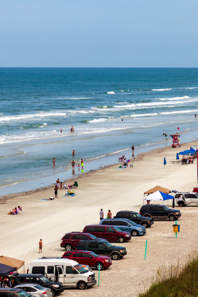 Beach scene with people walking and playing along the shoreline, cars parked directly on the sand, and tents set up for shade under a clear sky with gentle ocean waves in the background.
